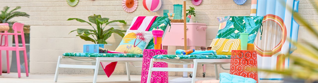 Lounge chairs with towels, pillows, colorful metal tables, pink umbrella, and colorful summer decorations.