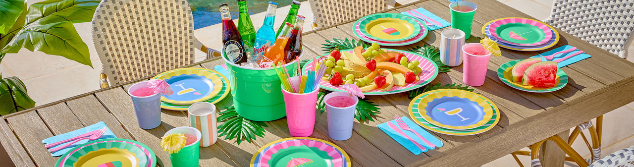 Poolside table set with colorful melamine dinnerware with stripes and summer icons, ice bucket, and matching drinkware.