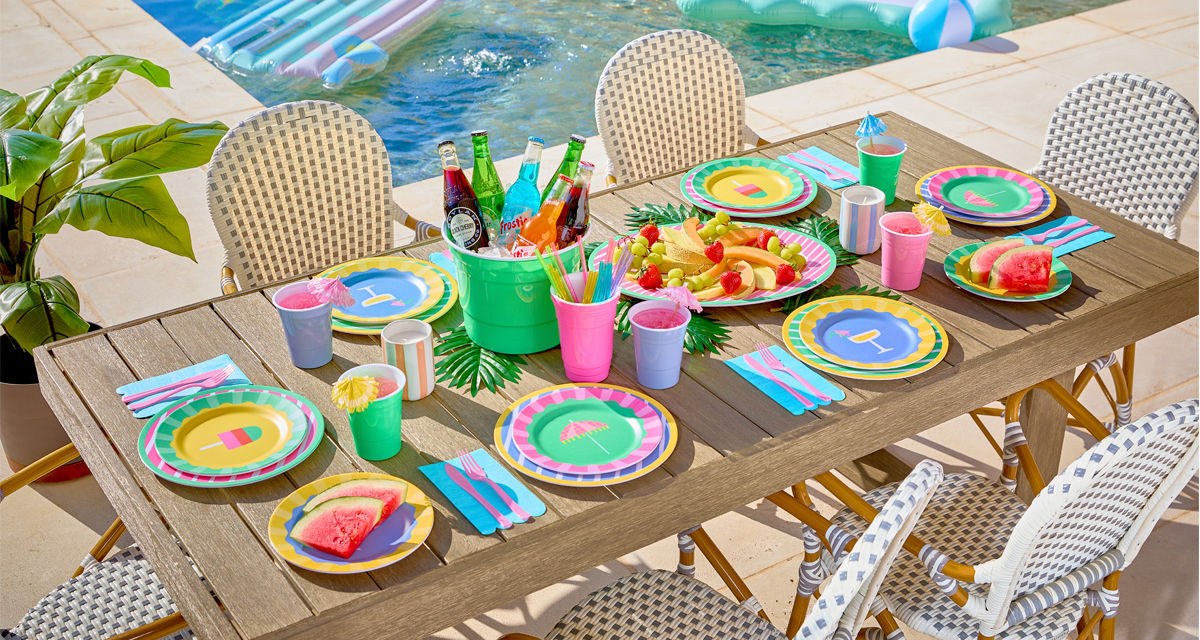 Poolside table set with colorful melamine dinnerware with stripes and summer icons, ice bucket, and matching drinkware.