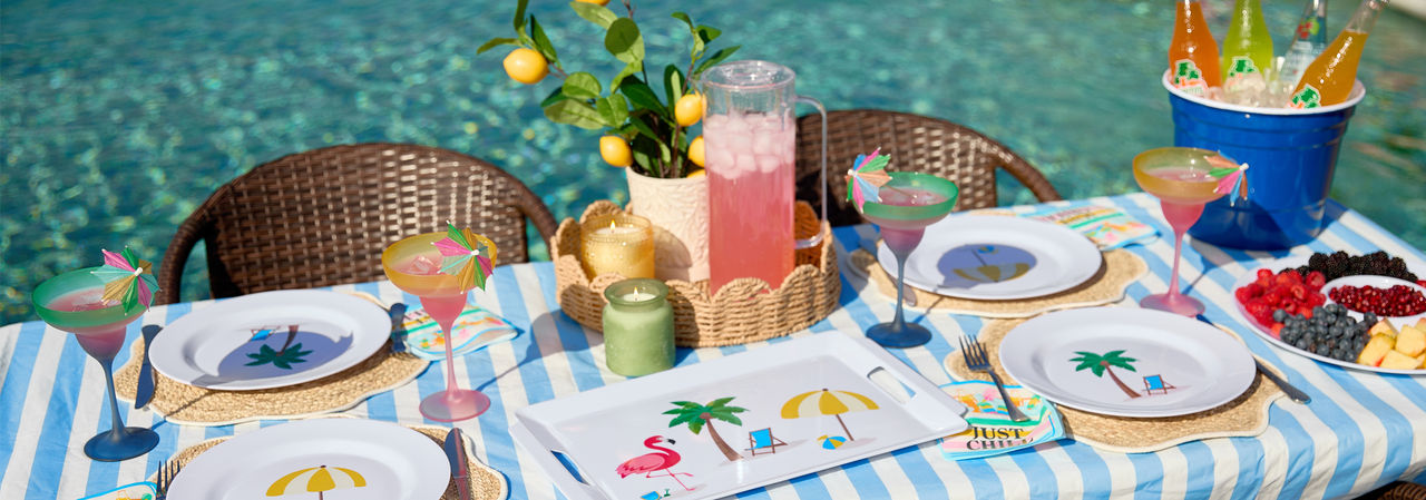 Poolside table set with colorful melamine dinnerware with summer icons, ice bucket, and plastic margarita glasses.