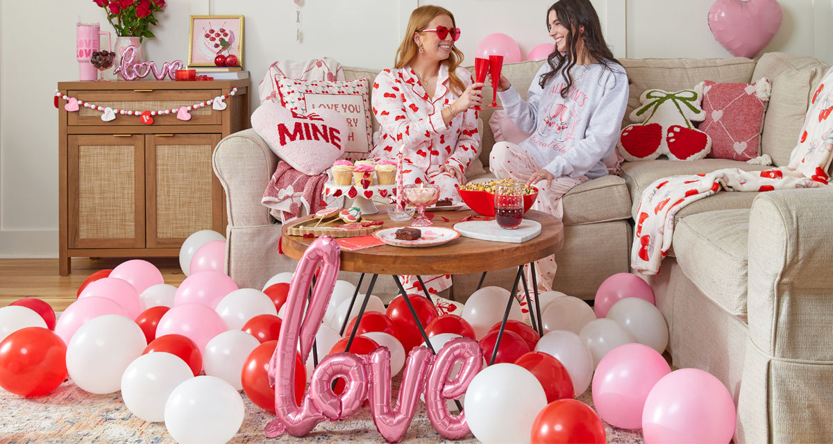 Galentine's Day living room setup, girls on couch toasting with heart-shaped plastic champagne glasses drinks, surrounded by Valentine's decor, balloons, pillows, throws, food, garland, banners, and more.