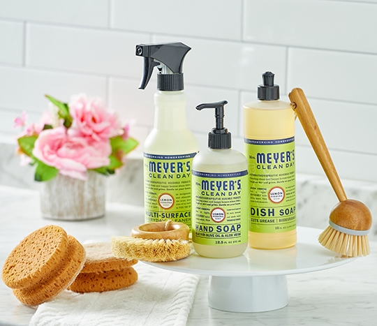 Mrs. Meyer cleaning products on stand surrounded by eco-friendly cleaning sponges and brushes on white counter top