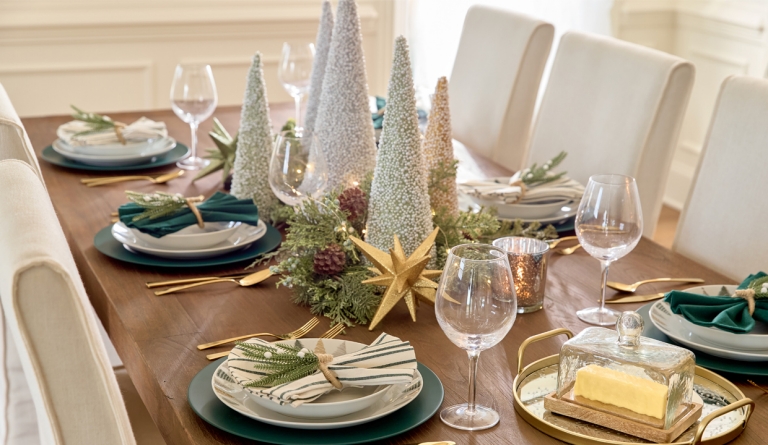 Dining table set for Christmas with ceramic Christmas tree designed dinner plates and bowls, wine glasses, solid and green-striped napkins with pine napkin holders, cone tree centerpiece with faux greenery, lights, and large sparkly tabletop decor.