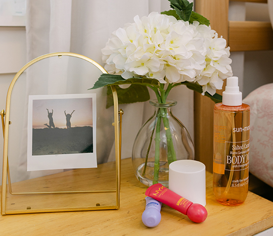A dorm bedside table is decorated with faux flowers, a glass vase, gold picture frame, and self-care college essentials from pOpshelf.