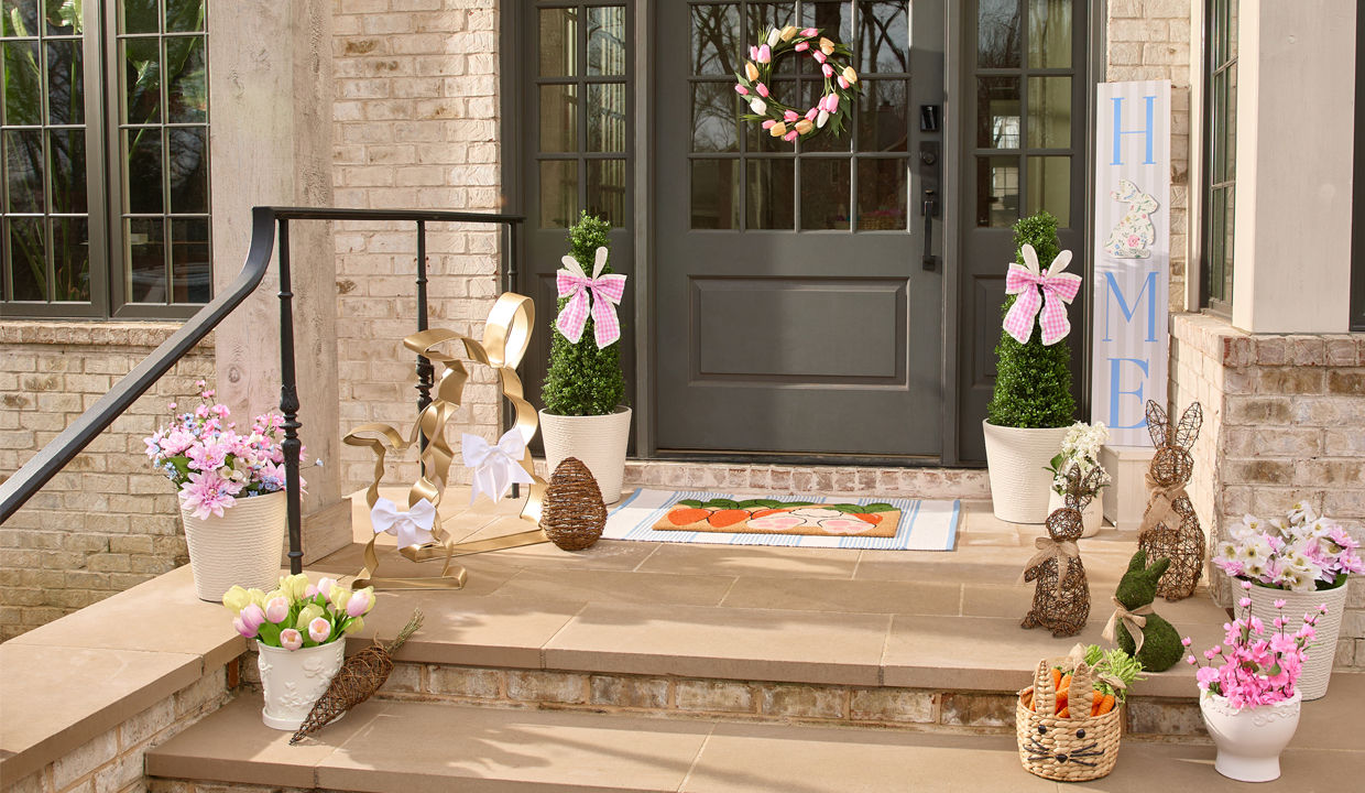 Front porch decorated for spring and easter with rattan floral baskets, tulip wreath, rattan bunnies & carrots, moss bunny, large metal bunnies, porch leaner, coir mat & more.