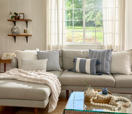 A bright white living room couch with a chaise lounge is covered with a soft white throw blanket and various blue plush pillows featuring stripes and shades of blue with decorated shelves above and a glass coffee table with a decorative tray display in front of it.