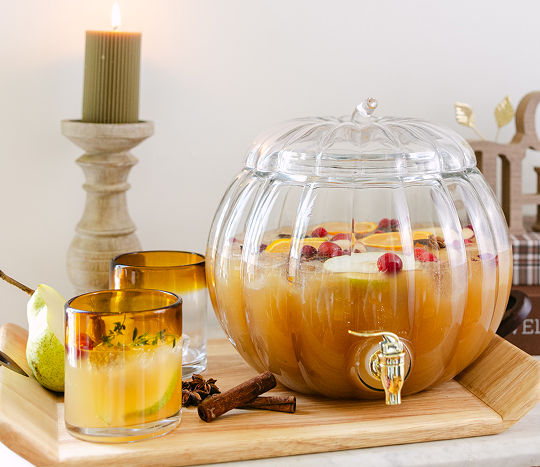 A clear pumpkin drink dispenser is filled with a fall drink garnished with cherries, oranges, and other fruit, then served in autumn drink glasses with orange rims sitting next to the autumn drink dispenser.
