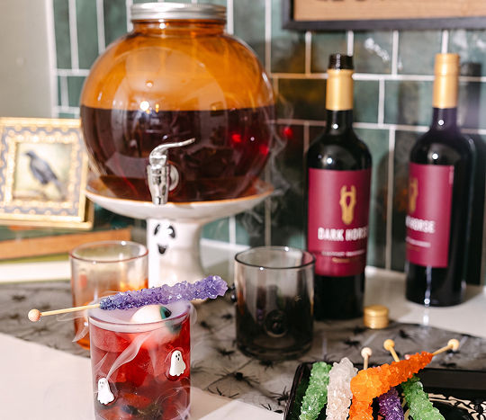 A kitchen counterspace is decorated with an orange drink dispenser for parties and featured sitting on top of a ghost-themed riser with pumpkin and Halloween icon glasses from pOpshelf.