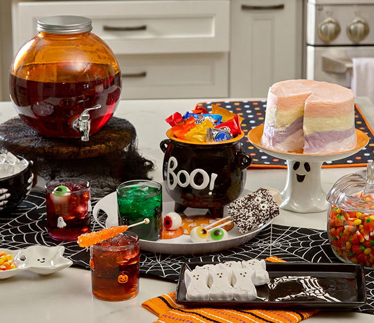 A kitchen island counter is decorated with classic Halloween-themed decor including a Boo! caldron, ghost cake display, spider web table runner, skeleton hand plates and bowls, an orange drink dispenser, and Halloween icon glassware.