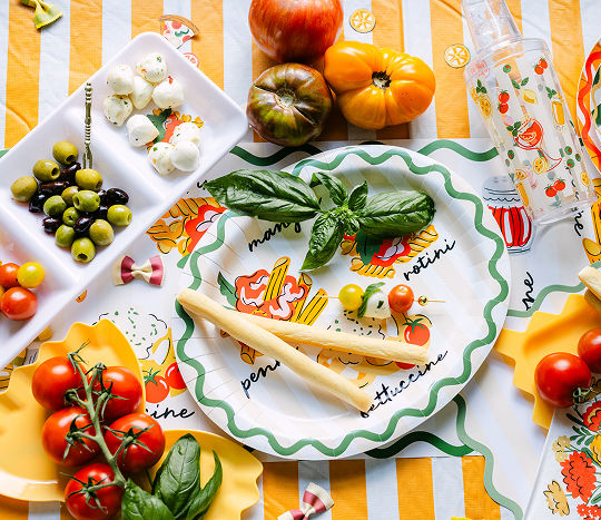 A vibrant yellow and white striped tablecover is set with colorful Italian noodle themed serveware that features green squiggles, a tray with various pasta shapes, pasta noodles-shaped serveware, and italian meal ingredients including basil, peppers, tomatos, olives, and garlic.