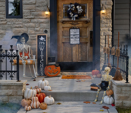 A stone home’s front porch is decorated for Halloween with affordable Halloween decor from pOpshelf, featuring full-sized decorative skeletons, a decorative candy cauldron, various pumpkin accessories, and a front porch leaner that says “It’s October Witches.”