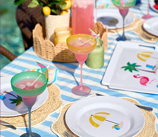 An outdoor dining table set with colorful vacation themed dinnerware, plastic gradient margarita glasses, and party supplies featuring tropical palm trees and vacation icons.