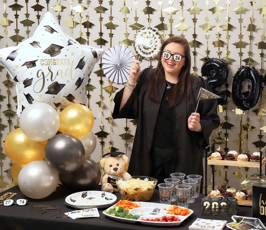 Grad in black gown at a grad party table featuring black & gold balloons, paper party supplies, foil decorations, photo booth props, gifts, and more from pOpshelf.
