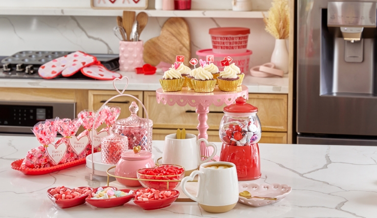 Kitchen island with Valentine's cake stand, trays, mugs, and candy with Valentine's Day kitchen linens , utensil holder, and baking containers in the background.