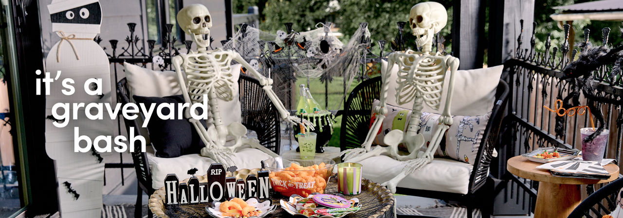 A porch is set with two faux skeletons sitting in lounge chairs with a table decorated with Halloween candy and snacks and decorated with fall florals and a classic Halloween Jack-O-Lantern themed pumpkin.