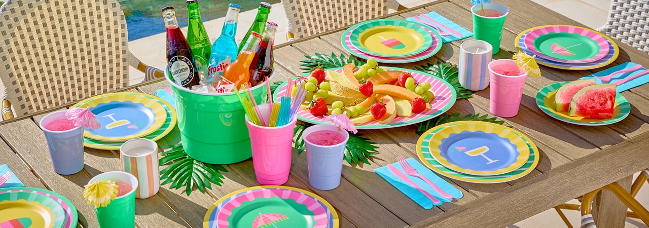Poolside table set with colorful melamine dinnerware with stripes and summer icons, ice bucket, and matching drinkware.