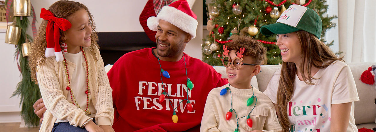 Family in front of Christmas tree wearing Santa hats, novelty Christmas glasses, Christmas bead necklaces, antler and tree headbands, and more.