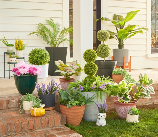 Various colors and designs of pOpshelf outdoor planters, flower pots, and plant stands filled with artificial greenery and flowers on a front porch.