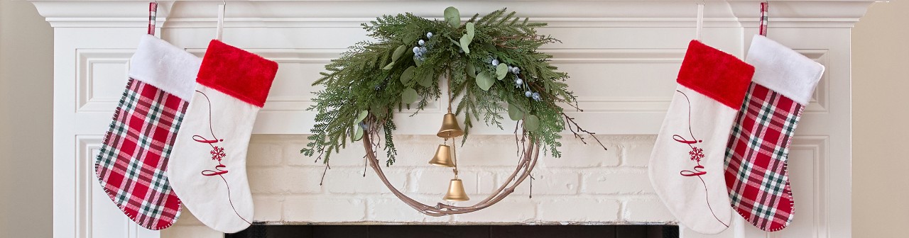 Red plaid and white 'Joy" stockings on a fireplace mantel decorated for Christmas with a wreath, large Christmas sign, trees, greenery & more.