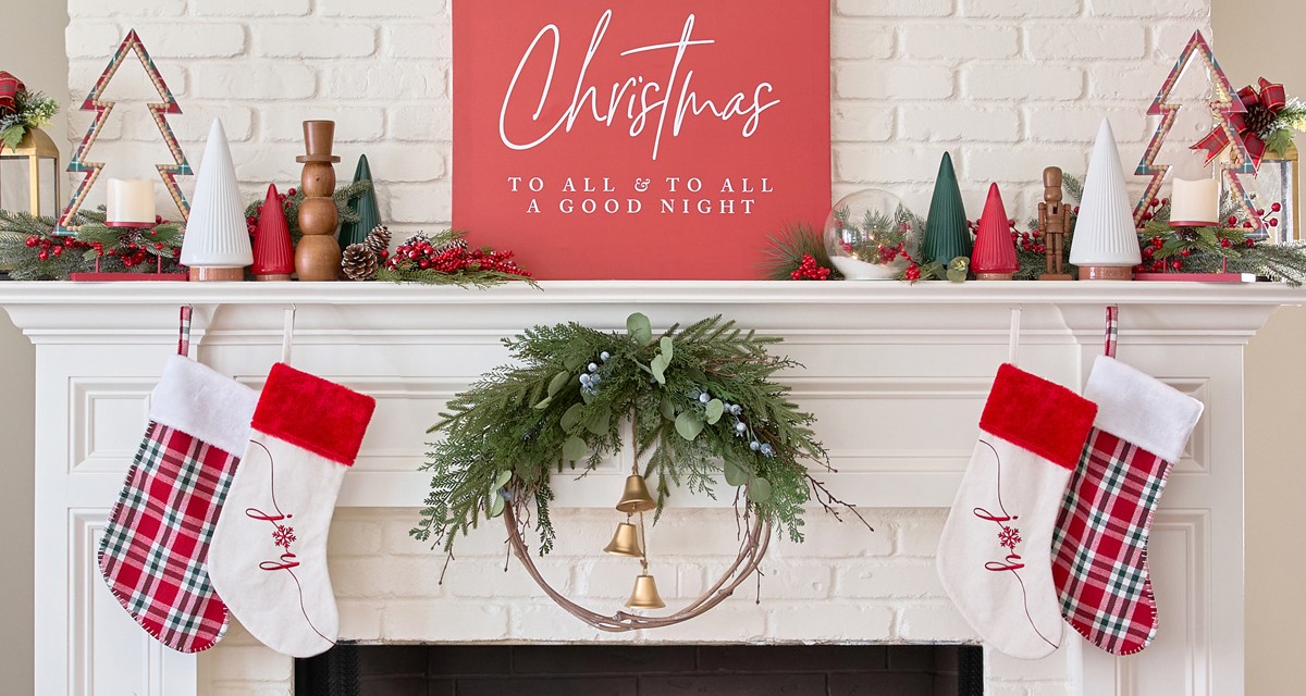 Red plaid and white 'Joy" stockings on a fireplace mantel decorated for Christmas with a wreath, large Christmas sign, trees, greenery & more.